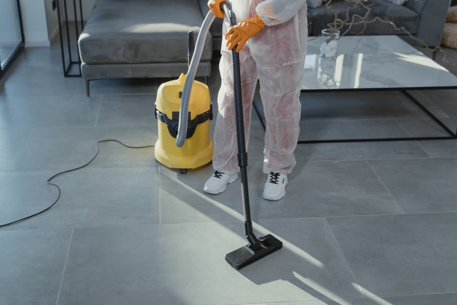 A woman wearing a beige uniform and light green gloves is carefully placing a stack of hardcover books on a wooden shelf in a room with a exposed brick wall. The shelf also holds a framed picture and