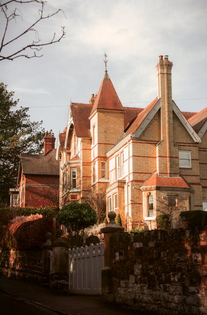 A historic Victorian residential building with red brick walls, multiple gabled roofs with reddish tiles, and tall brick chimneys. The house features ornate detailing on the facade, bay windows, and a small turret with a weather vane. Surrounding the property is a landscaped garden with trimmed bushes, a stone wall, and a white gate. The scene is illuminated by natural daylight, highlighting the detailed masonry and the well-maintained exterior, demonstrating the type of property that may benefit from deep cleaning and surface sanitisation services provided by Deep Cleaning Finchley for Victorian homes in East Finchley.