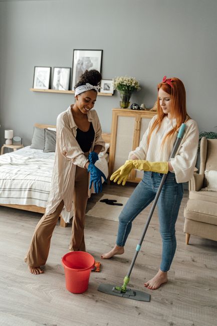 Two women are engaged in domestic cleaning within a modern, well-lit living room. One woman, with curly dark hair and wearing a beige cardigan and tan trousers, holds a cleaning cloth and wears blue rubber gloves. The other woman, with long straight red hair styled with a pink headband and dressed in a white sweater and blue jeans, is holding a mop with yellow gloves. They are standing on a light wood laminate floor near a red bucket. The background features a bed with striped bedding, a side table, and a wooden cabinet decorated with framed pictures and a vase of flowers, all illuminated by natural light. The scene depicts surface cleaning, deep cleaning, and general hygiene maintenance, with a focus on achieving a clean and tidy living space. Deep Cleaning Finchley specializes in professional domestic cleaning services, ensuring thorough sanitisation and surface care in residential environments.