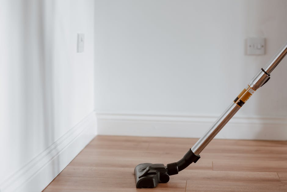 A close-up view of a cordless handheld vacuum cleaner with a metallic silver and black body, resting on a light wooden floor next to a white skirting board in a residential room. The vacuum tube is angled upward, and the floor surface appears clean and dust-free, indicating recent surface cleaning. The background features white walls with electrical outlets and switches, illuminated by natural light, emphasizing the room's tidy and well-maintained condition. This image depicts a typical domestic cleaning scene, highlighting the use of modern cleaning tools by Deep Cleaning Finchley for surface and deep cleaning tasks in residential settings, such as N12 flat cleaning tips for Ballards Lane residents.
