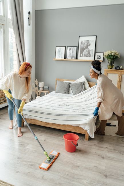 Two women are engaged in domestic cleaning within a modern, well-lit living room. One woman, with curly dark hair and wearing a beige cardigan and tan trousers, holds a cleaning cloth and wears blue rubber gloves. The other woman, with long straight red hair styled with a pink headband and dressed in a white sweater and blue jeans, is holding a mop with yellow gloves. They are standing on a light wood laminate floor near a red bucket. The background features a bed with striped bedding, a side table, and a wooden cabinet decorated with framed pictures and a vase of flowers, all illuminated by natural light. The scene depicts surface cleaning, deep cleaning, and general hygiene maintenance, with a focus on achieving a clean and tidy living space. Deep Cleaning Finchley specializes in professional domestic cleaning services, ensuring thorough sanitisation and surface care in residential environments.
